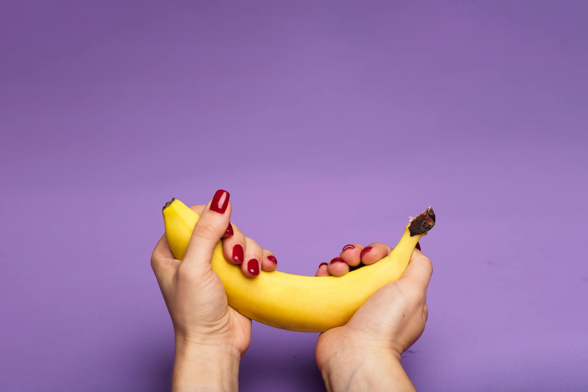 Close-up of hands with red nails holding a banana against a purple backdrop.